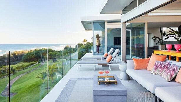 A modern outdoor seating area with a view of the ocean, a glass balustrade, and a sofa with cushions. - Beachfront Hideaway (Sunrise Beach)