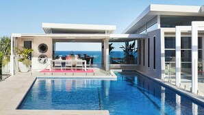 A modern pool area with a clear view of the ocean, a covered seating area, and a glass-enclosed structure. - Beachfront Hideaway (Sunrise Beach)