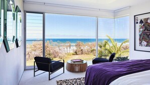 A modern bedroom with a large window overlooking a beach, a bed, two chairs, and a small round table. - Beachfront Hideaway (Sunrise Beach)