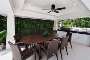 A modern dining area with a round table, wicker chairs, a ceiling fan, and a kitchen area with a sink and countertop.