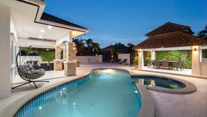 A swimming pool with a curved design, surrounded by a covered patio area with seating and a small table.