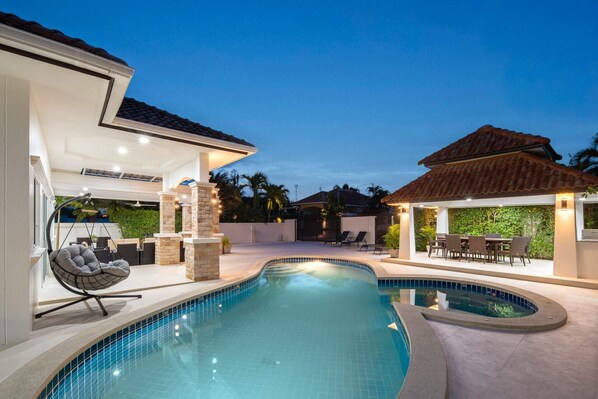 A swimming pool with a curved design, surrounded by a covered patio area with seating and a small table.