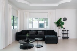 Un salon moderne avec un canapé d’angle, deux tables rondes, une plante en pot et une horloge murale.