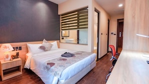 A modern hotel room with a large bed, a bedside table with a lamp, a mirror, and a desk with a chair.