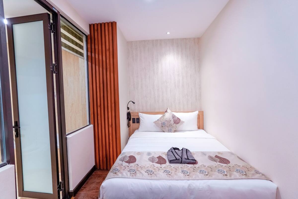 Habitación de hotel con cama, ventana con persianas, cabecero de madera y mesita de noche con lámpara.
