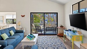 A living room with a sofa, coffee table, and a view through a sliding glass door to an outdoor balcony. - The Bungalow - Captiva Beach Cottage on Turner Beach (Captiva)