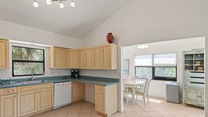 A kitchen with wooden cabinets, a dishwasher, a coffee maker, and a dining area with a table and chairs. - The Bungalow - Captiva Beach Cottage on Turner Beach (Captiva)