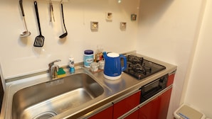 A kitchen with stainless steel sinks, red cabinets, a blue kettle, and various utensils hanging on the wall. - IBASHO Shibuya Higashi 604 (Tokyo)