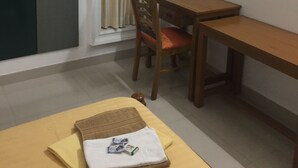 Ein kleiner, kompakter Raum mit einem Bett, einem Schreibtisch und einem Stuhl. Es gibt ein Fenster mit Vorhängen, eine Tür und ein Bild an der Wand.