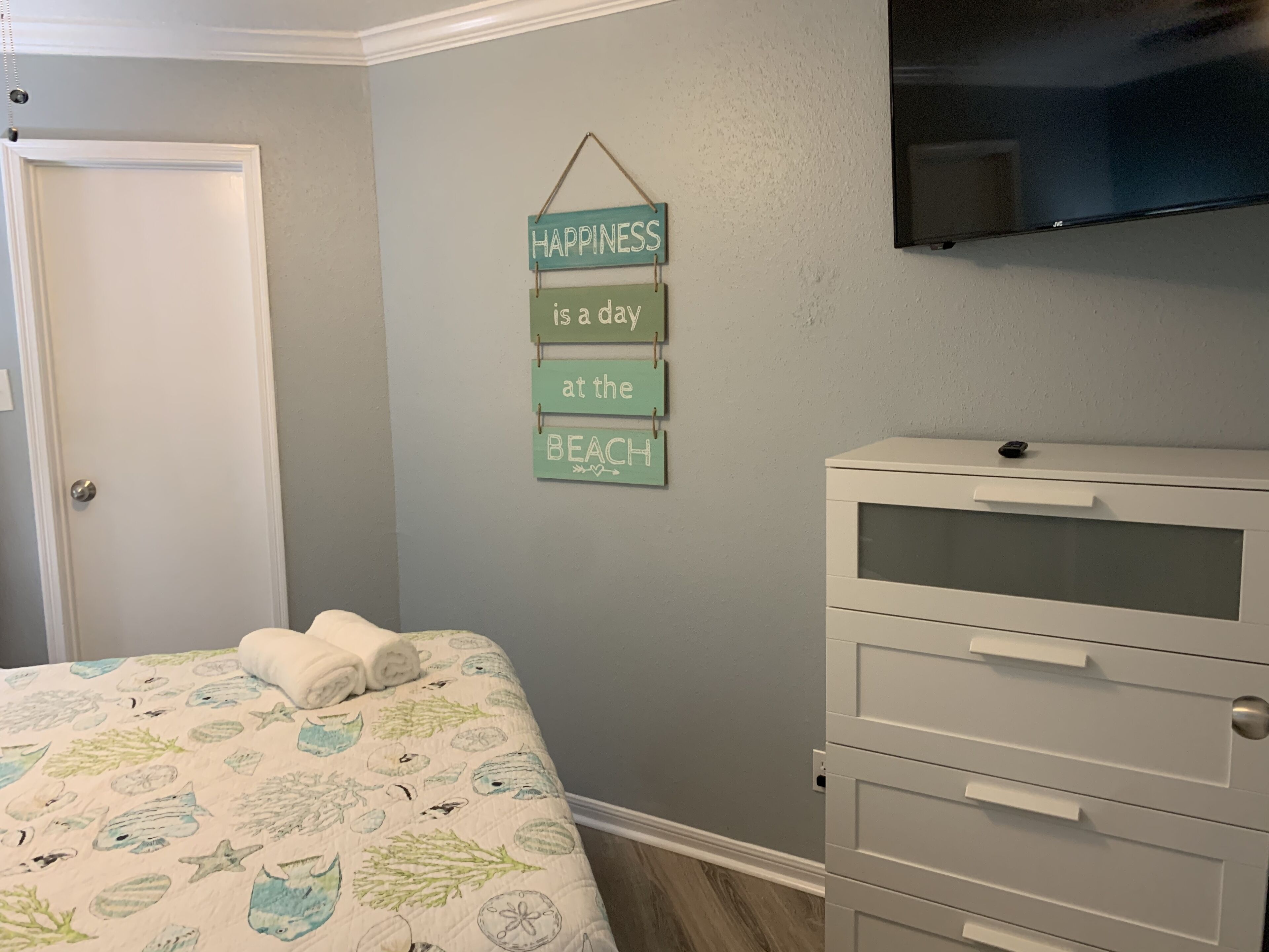 A bedroom with a bed, a dresser, and a wall-mounted sign that reads "HAPPINESS is a day at the beach."