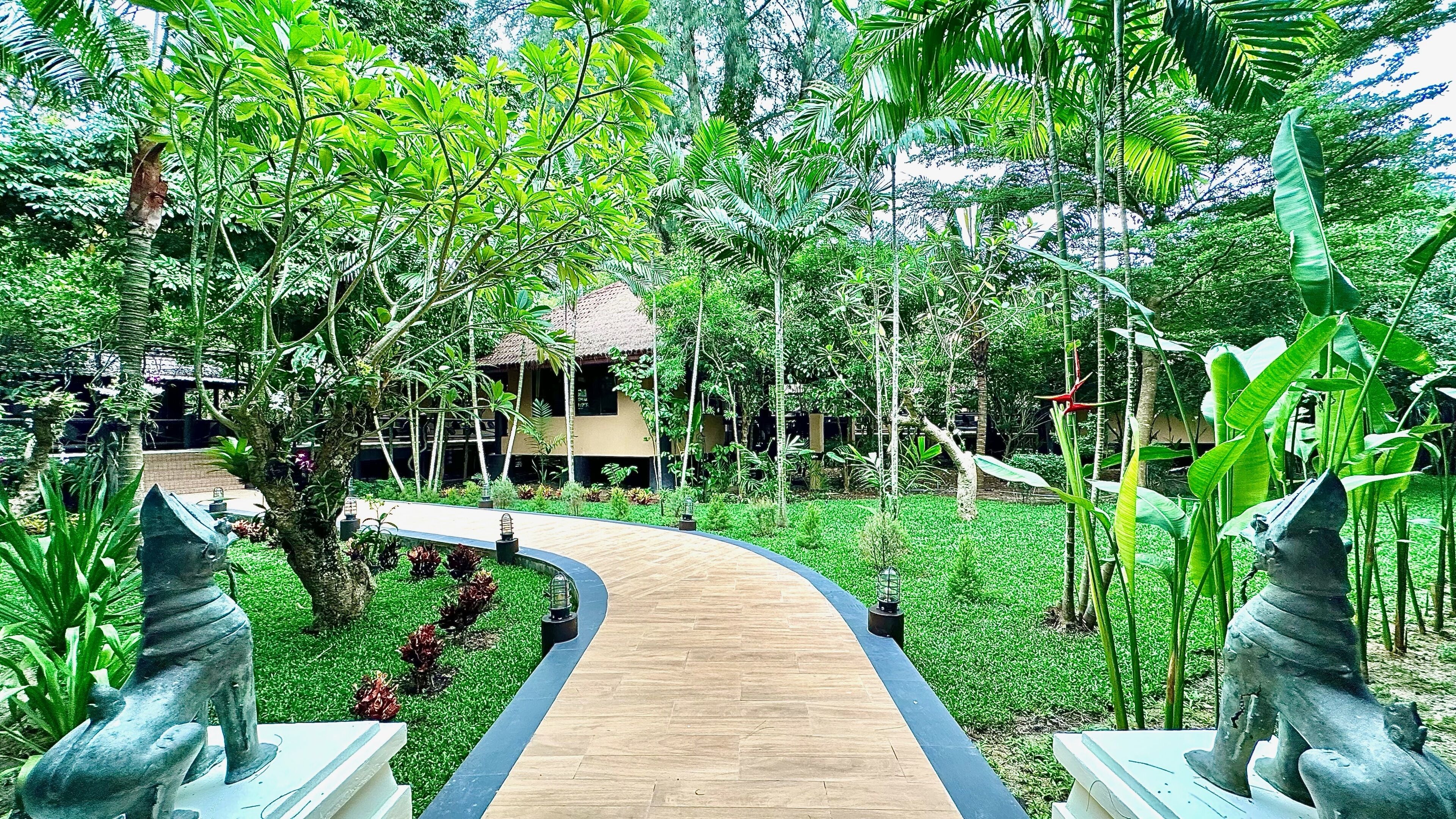 A wooden pathway leading to a building surrounded by lush greenery and tropical plants.