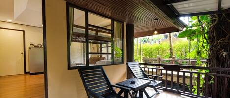 A balcony with wooden flooring, two black lounge chairs, and a view of greenery and a building in the background.