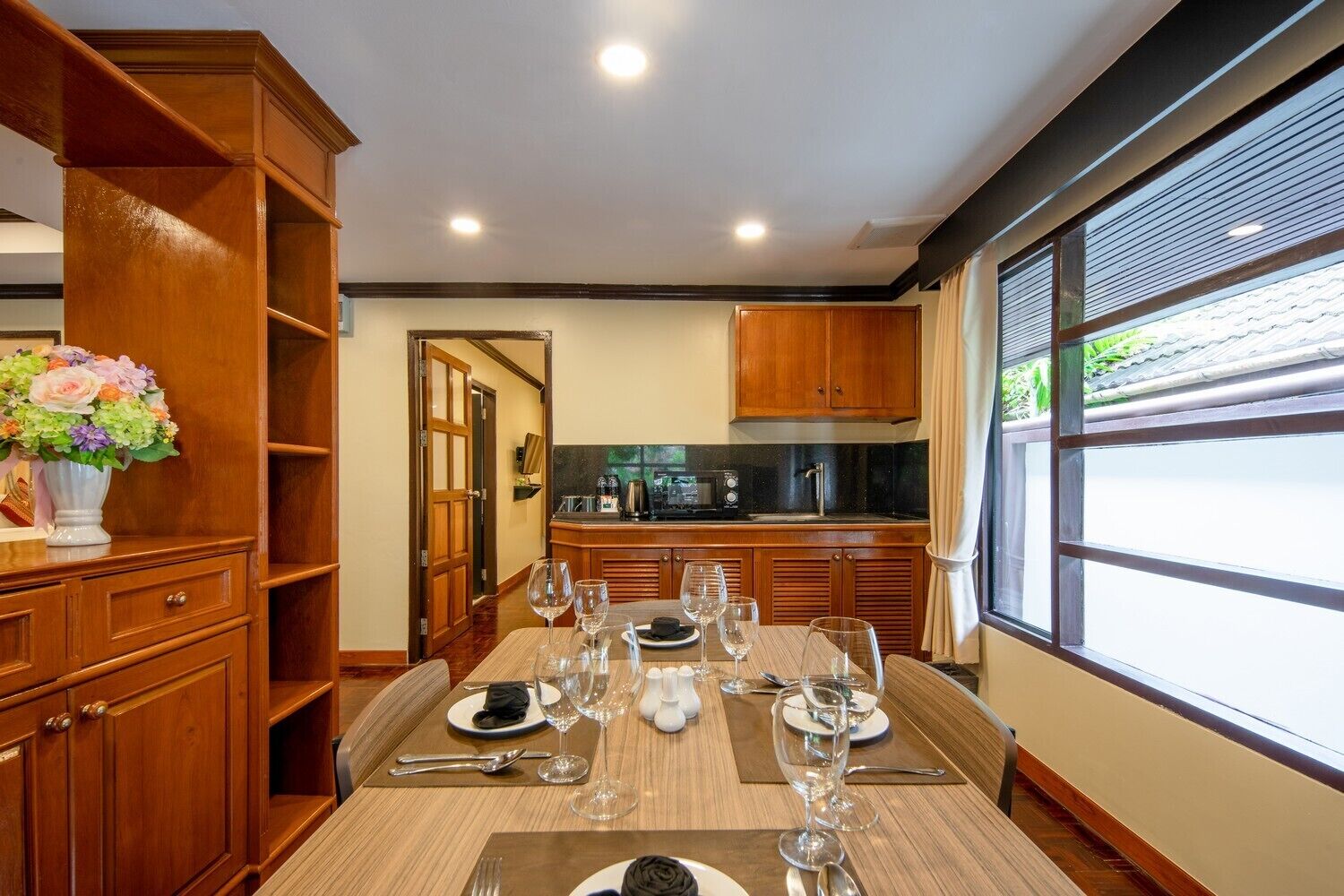 A dining area with a wooden table set for a meal, a kitchen in the background, and a large window with blinds.