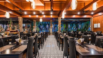 A restaurant with multiple tables and chairs, wooden ceiling beams, and hanging light fixtures.