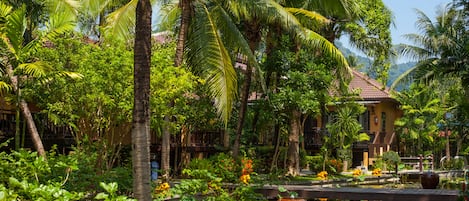A tropical garden with palm trees, a pond, and a building with a tiled roof.