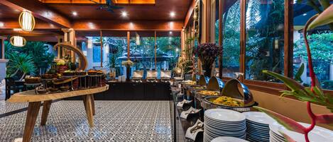 A buffet area with a variety of food items, a long table with plates, and a view of the outdoors through large windows.