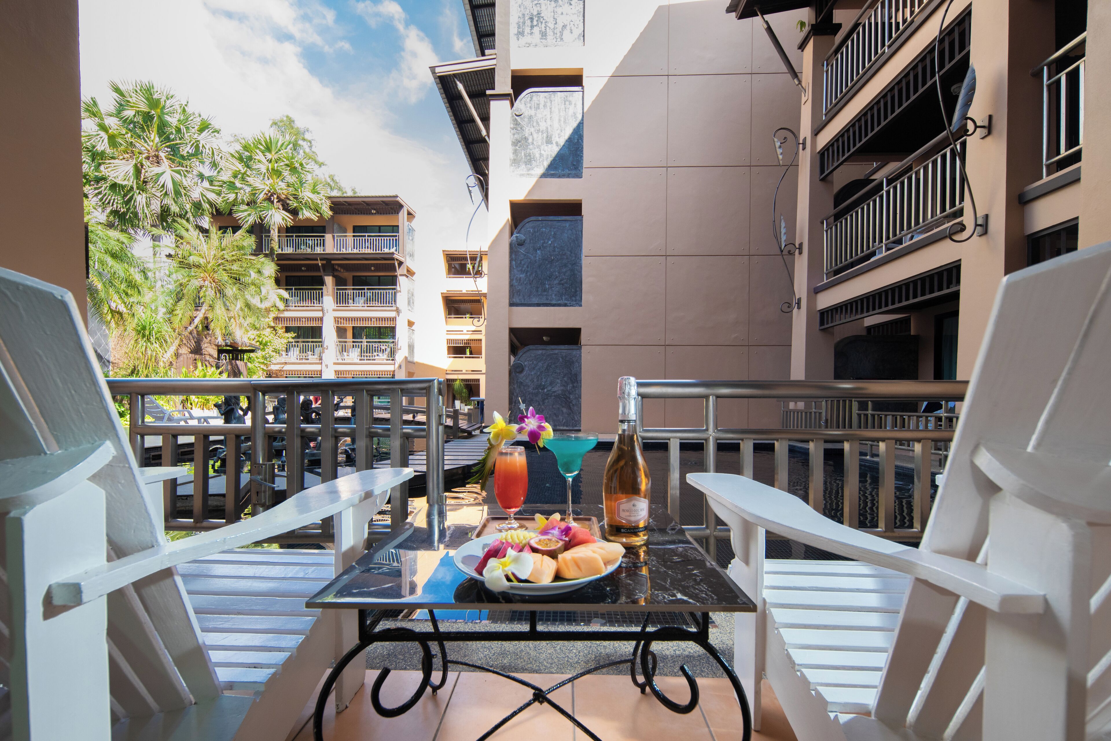 A balcony with a table set for breakfast, including a bottle of wine, fruit, and a drink.