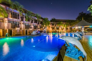 Una piscina con una escultura de delfín, palmeras y un edificio con balcones e iluminación cálida.