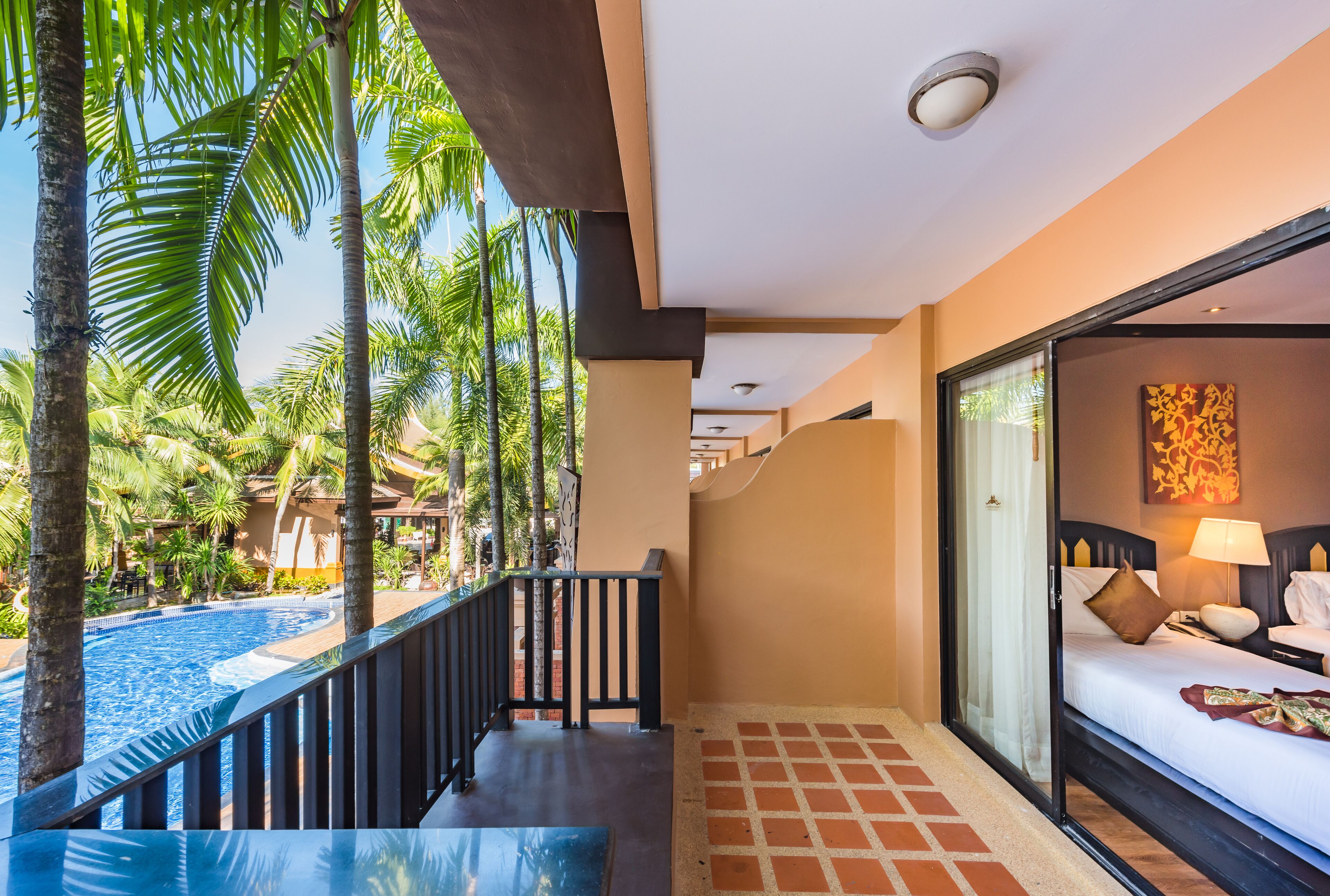 A hotel room with a view of a swimming pool, a balcony with a railing, and a bed with pillows and a lamp.