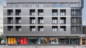 A modern multi-story hotel with a flat facade, multiple balconies, and a variety of ground-level retail spaces. - Aspira Gems Center Silom (Bangkok)
