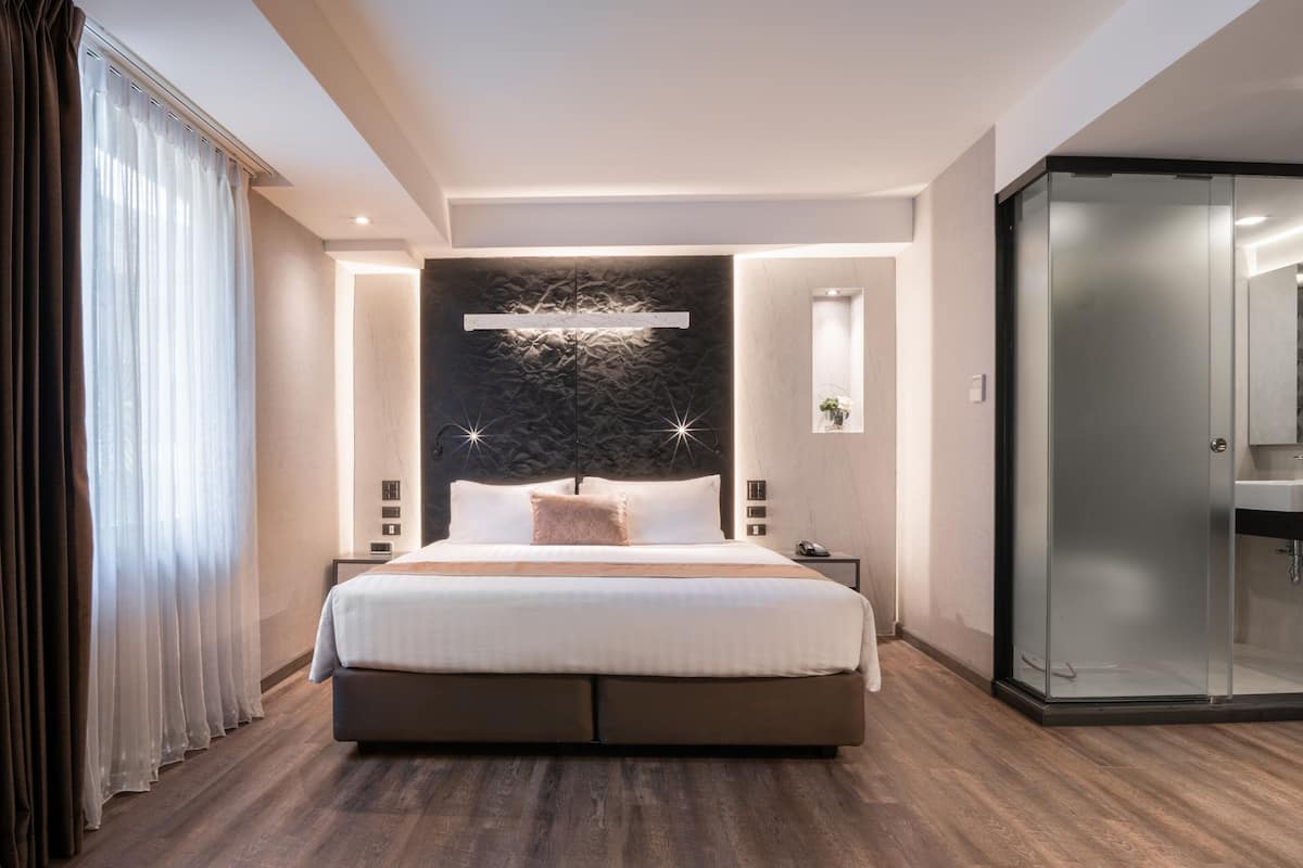 A modern hotel bedroom with a large bed, bedside tables, a glass shower enclosure, and a stone accent wall.