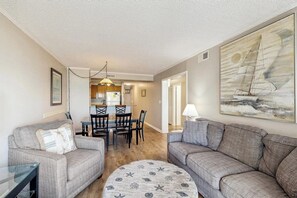 A living room with two grey sofas, a round coffee table with a nautical pattern, a dining area with a wooden table and chairs, and a large sailboat painting on the wall. - Cute and cozy condo with ocean view, pool & close to many attractions (Myrtle Beach)
