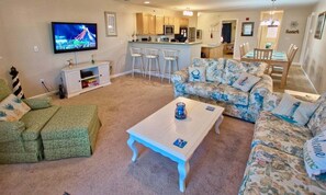 A living room with a TV, sofa, coffee table, and a kitchen area in the background.