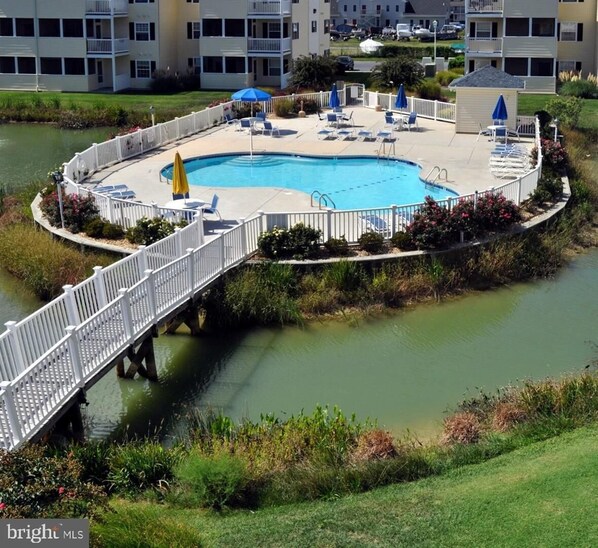 A swimming pool with a white deck, surrounded by lounge chairs and umbrellas, connected to a bridge over a body of water.