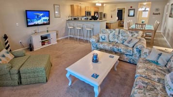 A living room with a TV, a sofa, a coffee table, and a kitchen area in the background.