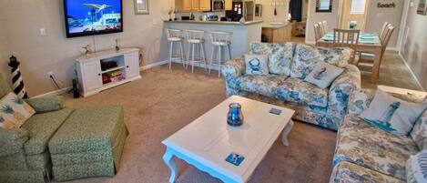 A living room with a TV, a sofa, a coffee table, and a kitchen area in the background.