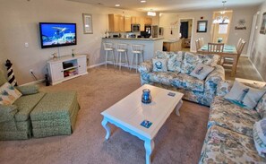 A living room with a TV, a sofa, a coffee table, and a kitchen area in the background. - The Palms * Luxury 3BR Condo End-Unit Home (Rehoboth Beach)