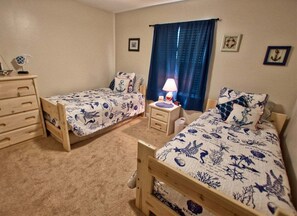 Een kamer met twee houten bedden, een nachtkastje met lamp, een dressoir en een raam met gordijnen.