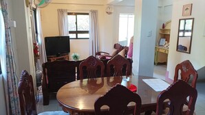 A dining area with a round wooden table and six chairs, a television on a stand, and a window with curtains.