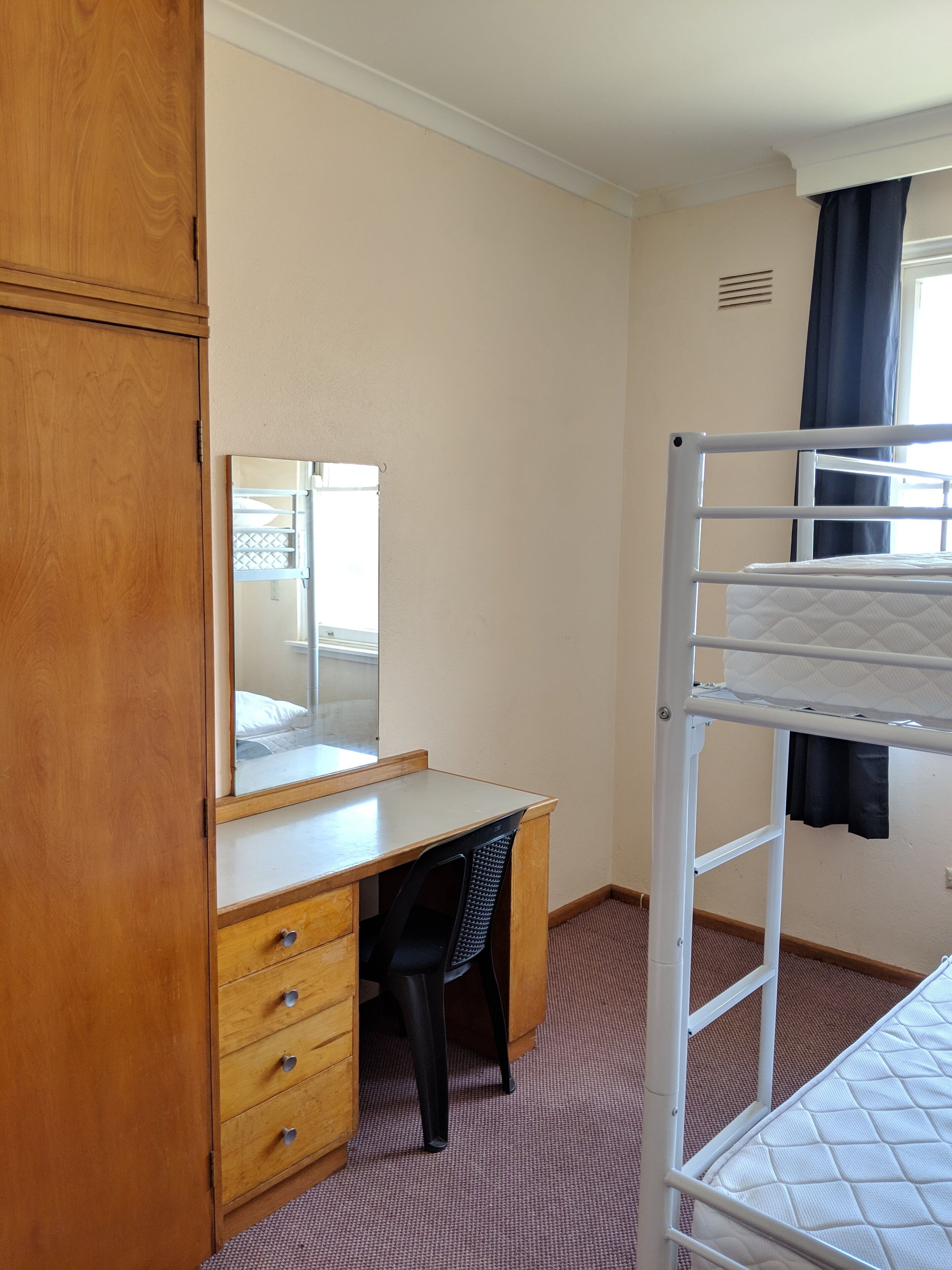 Une chambre avec un lit superposé, un bureau en bois avec des tiroirs, une chaise, un miroir et une fenêtre avec des rideaux.