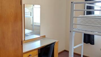 Une chambre avec un lit superposé, un bureau en bois avec des tiroirs, une chaise, un miroir et une fenêtre avec des rideaux.