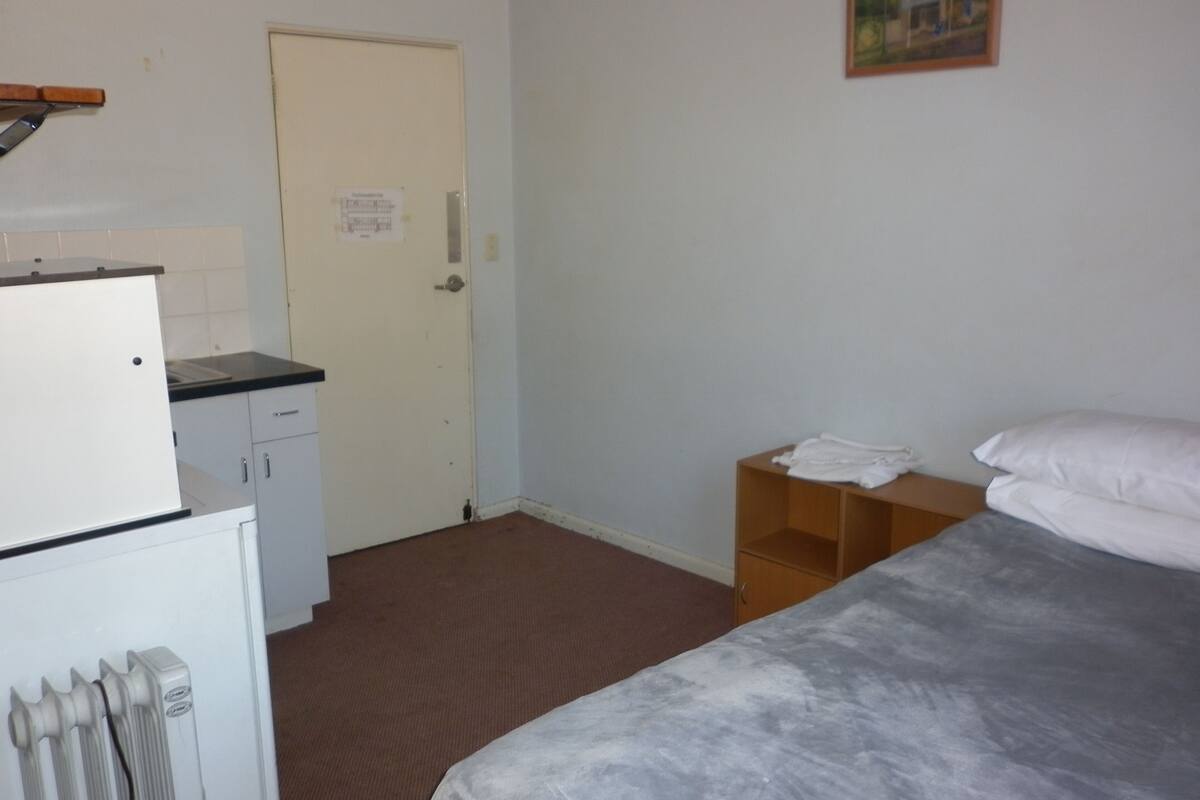 Una habitación pequeña y poco amueblada con una cama, una mesita, un radiador y una puerta con un aviso.