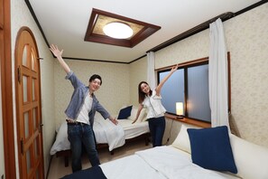 A hotel room with two beds, a wooden door, a ceiling light fixture, and a window with curtains.