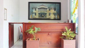 A reception area with a framed painting of a building, a wooden bench, and two potted plants. - Wisma Rajawali Cirebon (Cirebon)