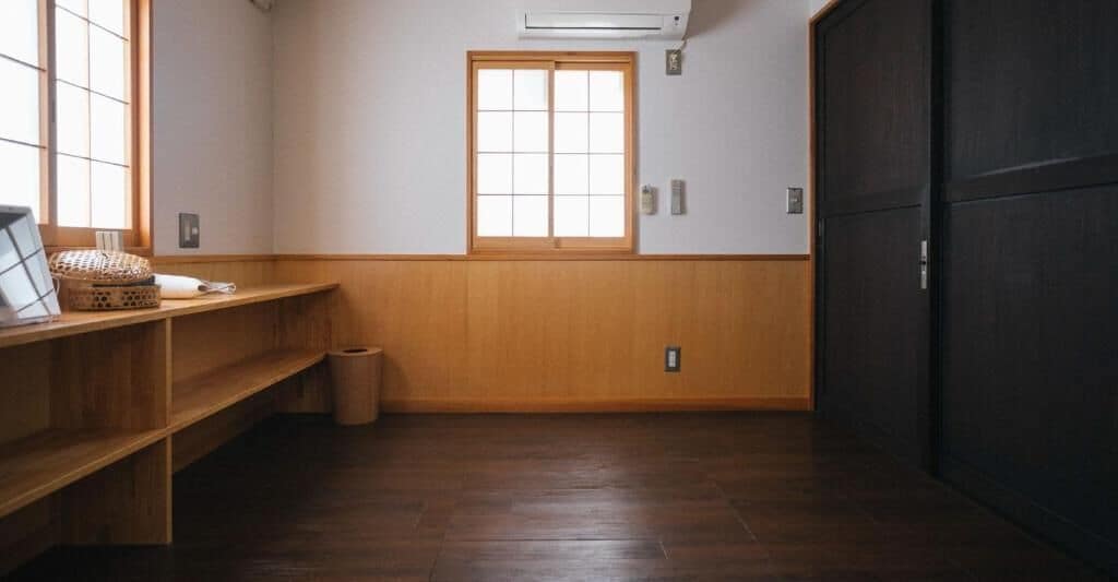 A room with a wooden floor, a window with a view, a shelf with items, and a door.
