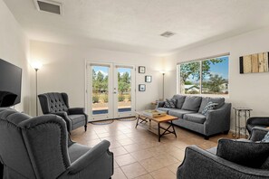 A living room with a grey sofa, armchairs, a coffee table, and a TV. There are two doors leading outside, and a window with a view of trees and a house. - Rustic Cottage Feel, Close to Wineries, Sleeps 6! (Cornville)