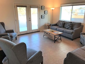 A living room with a sofa, chairs, a coffee table, and a lamp. There are framed pictures on the wall and a door leading outside.