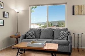 Sala de estar com sofá, mesa de centro, abajur e janela com vista para casas e colinas.