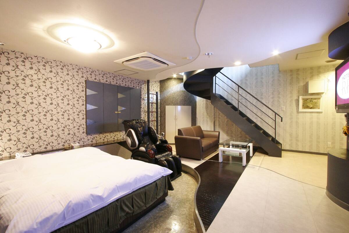 A modern hotel room with a bed, massage chair, and a staircase leading to an upper level.