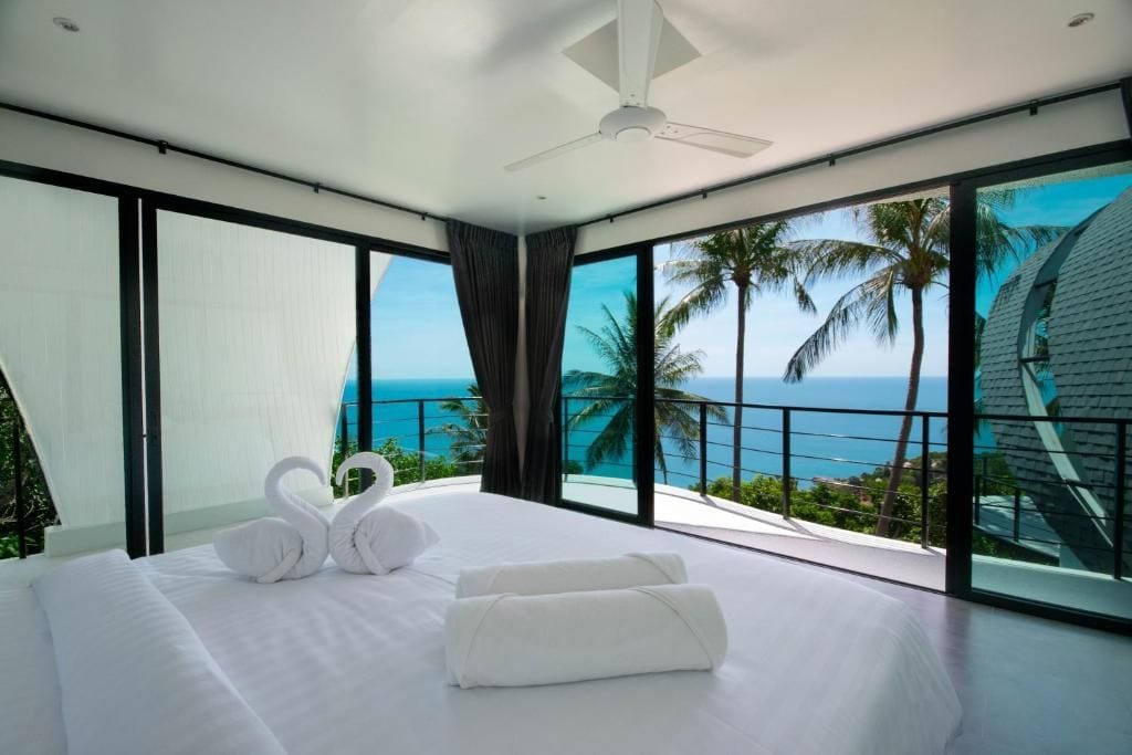Un dormitorio moderno con una cama grande, dos toallas con forma de cisne y vistas al océano y palmeras.