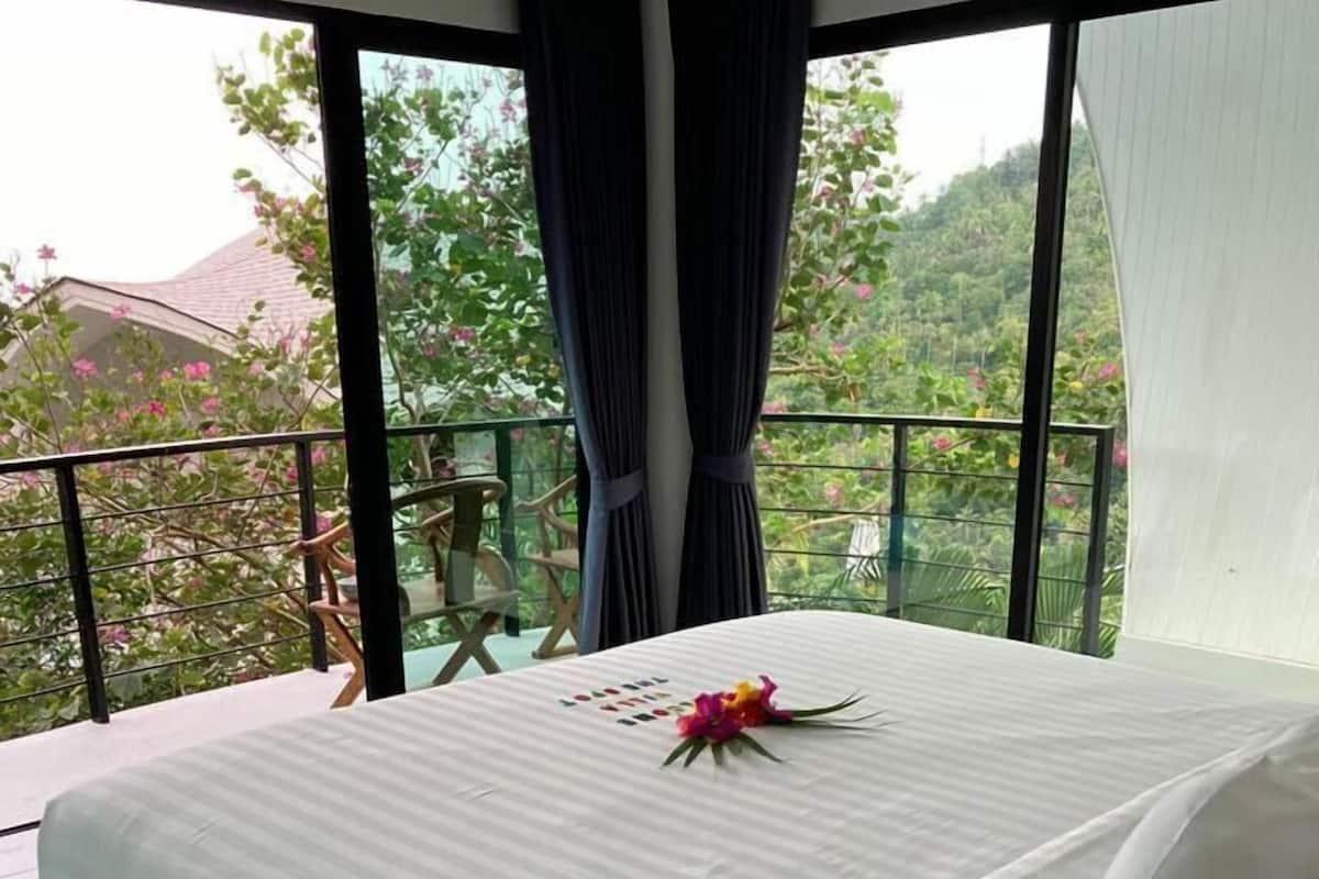 Quarto com cama grande, varanda com vista para vegetação e uma cadeira.