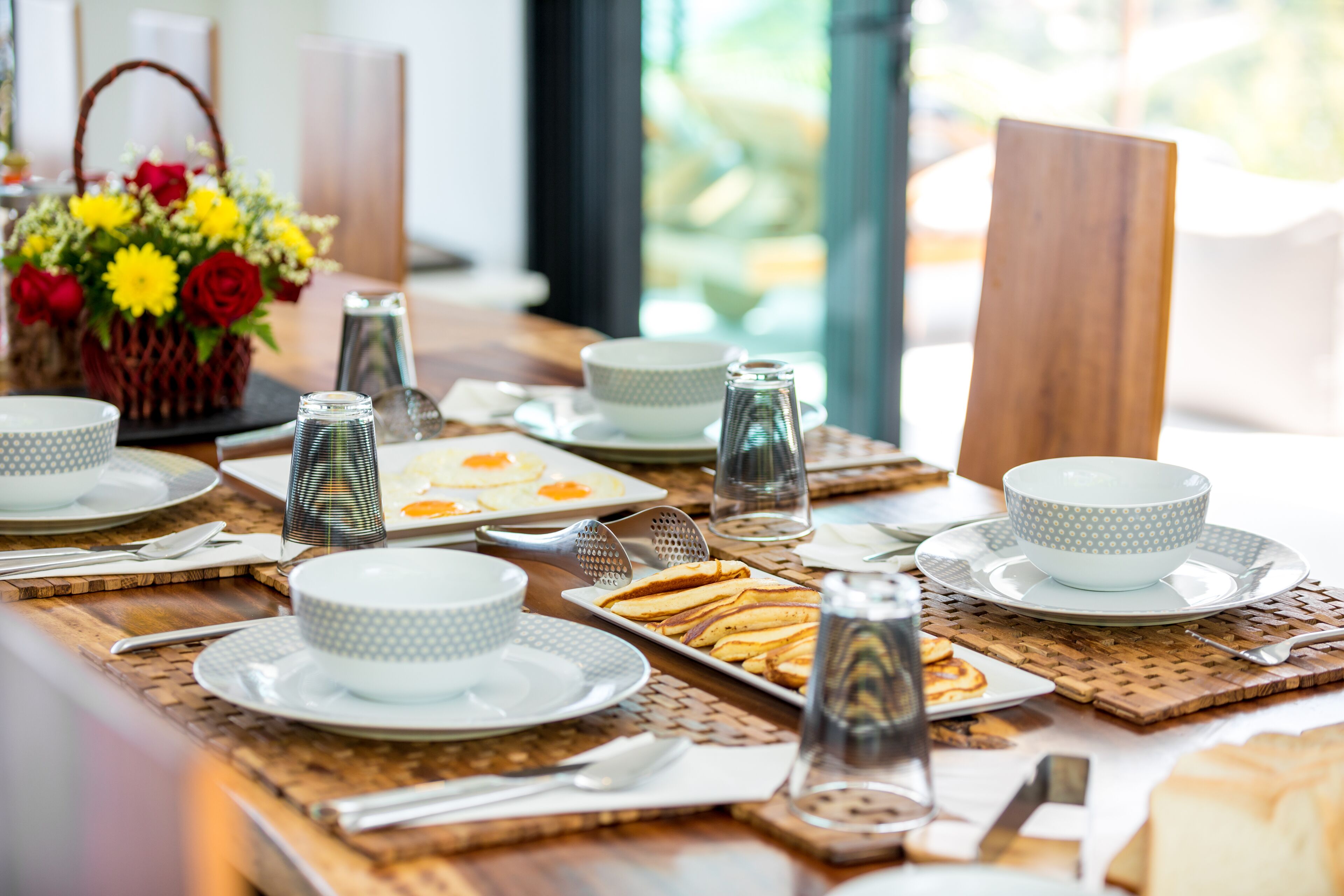 Una escena de desayuno con una mesa lista para dos personas, con platos, tazones, tazas y una cesta de flores.