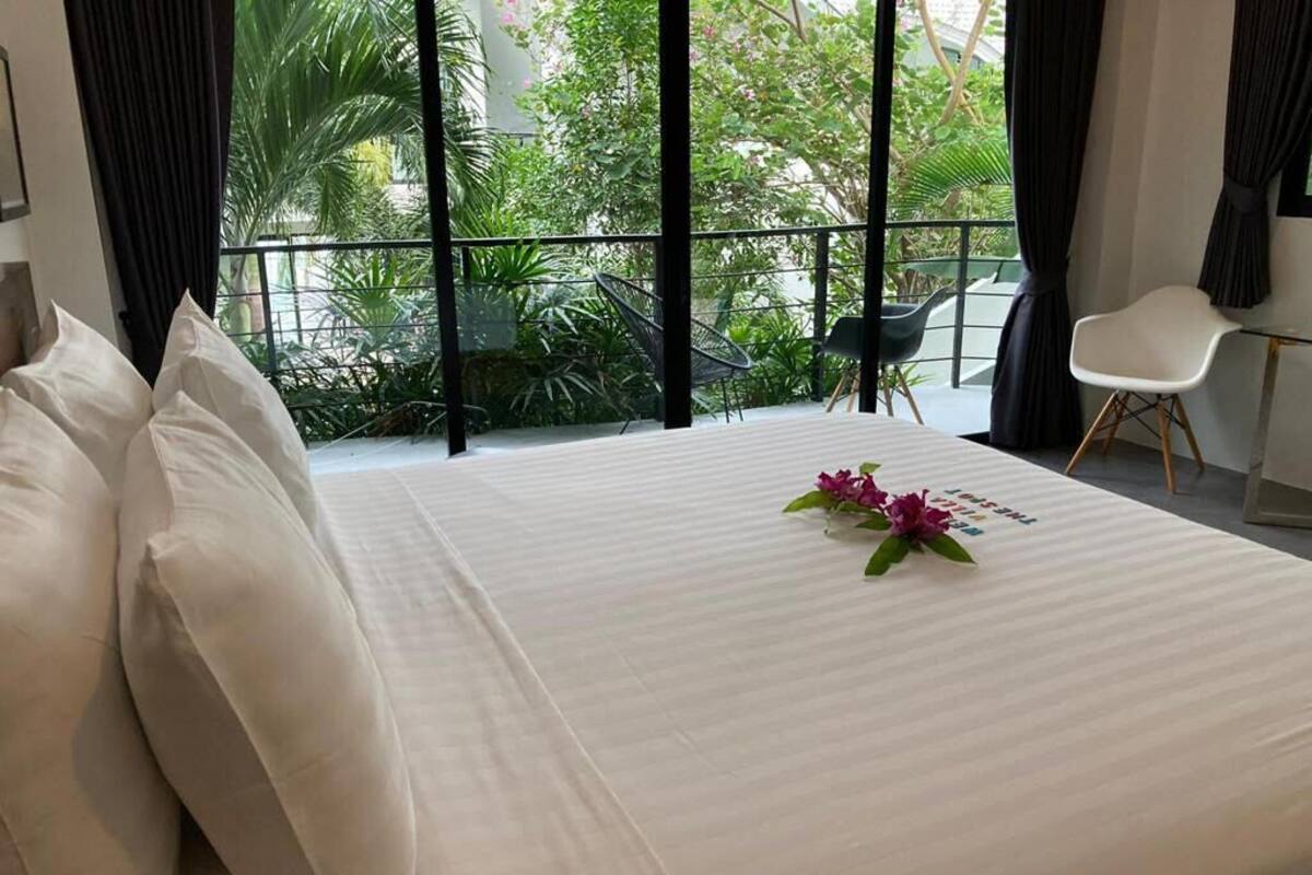 Quarto de hotel com uma cama grande, uma mesa pequena, uma cadeira e uma varanda com plantas e vista para vegetação.