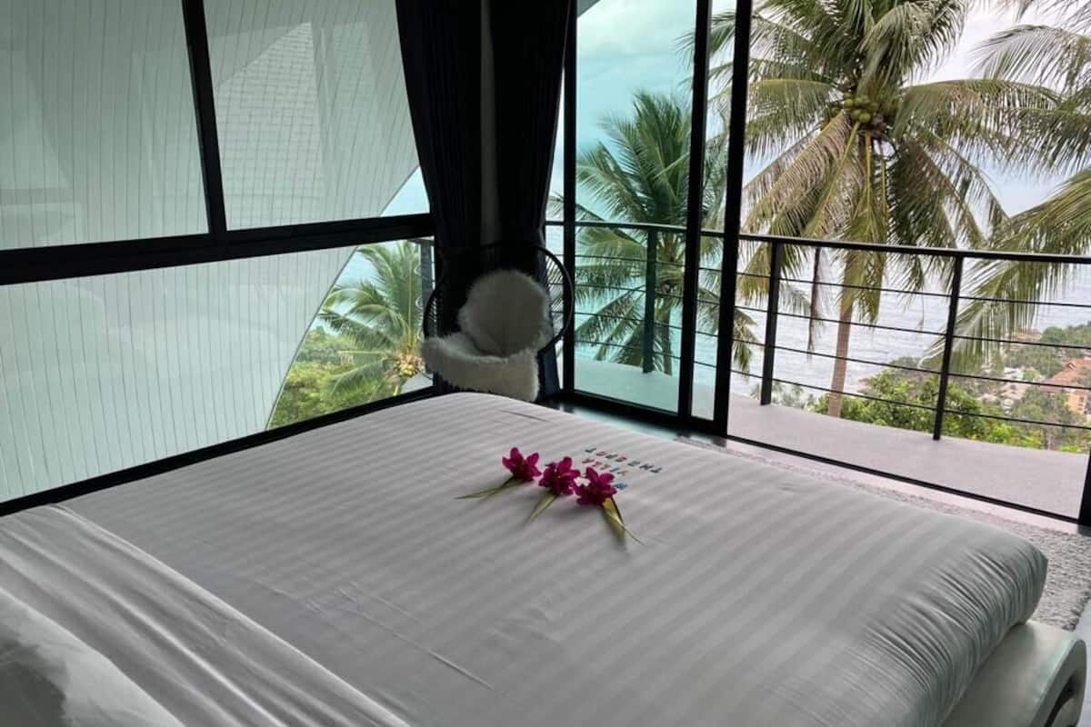 Quarto de hotel com uma janela grande, cama com roupa de cama branca e vista para palmeiras e uma praia.