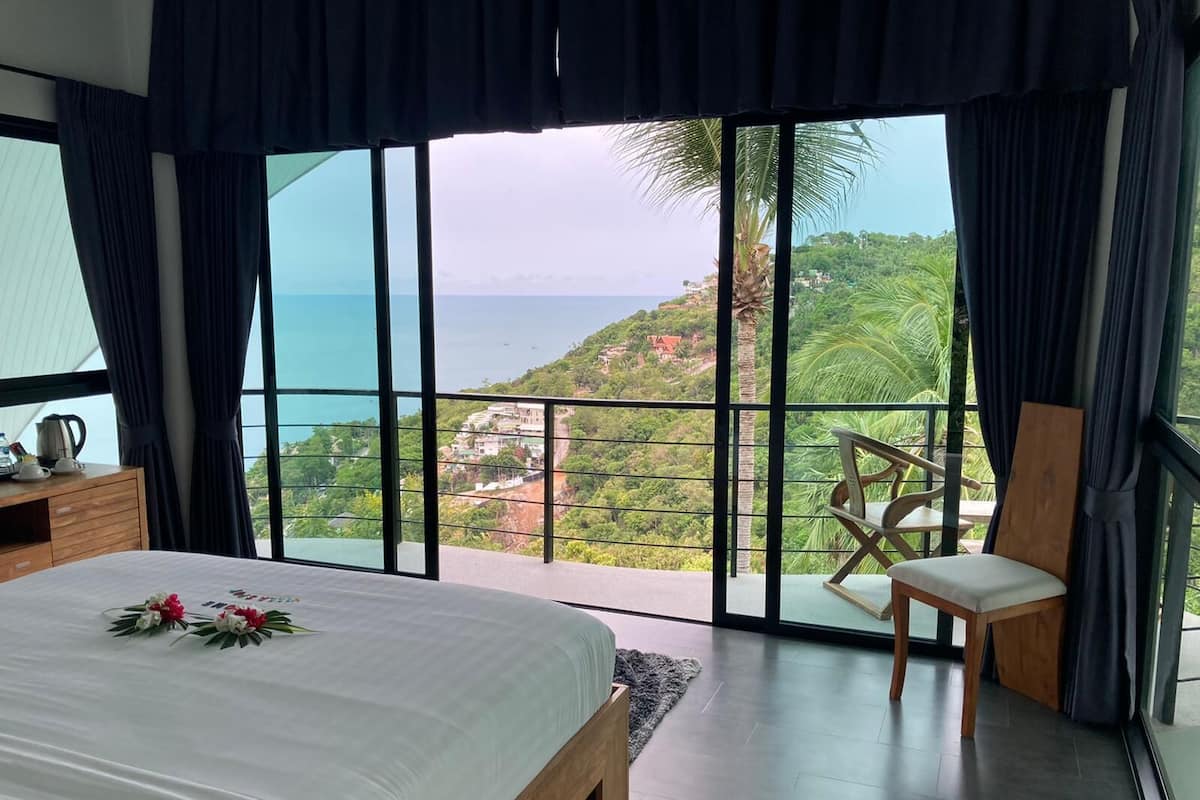 Quarto com uma cama grande, uma mesa de cabeceira de madeira, uma cadeira e vista para a paisagem costeira através de janelas do chão ao teto.