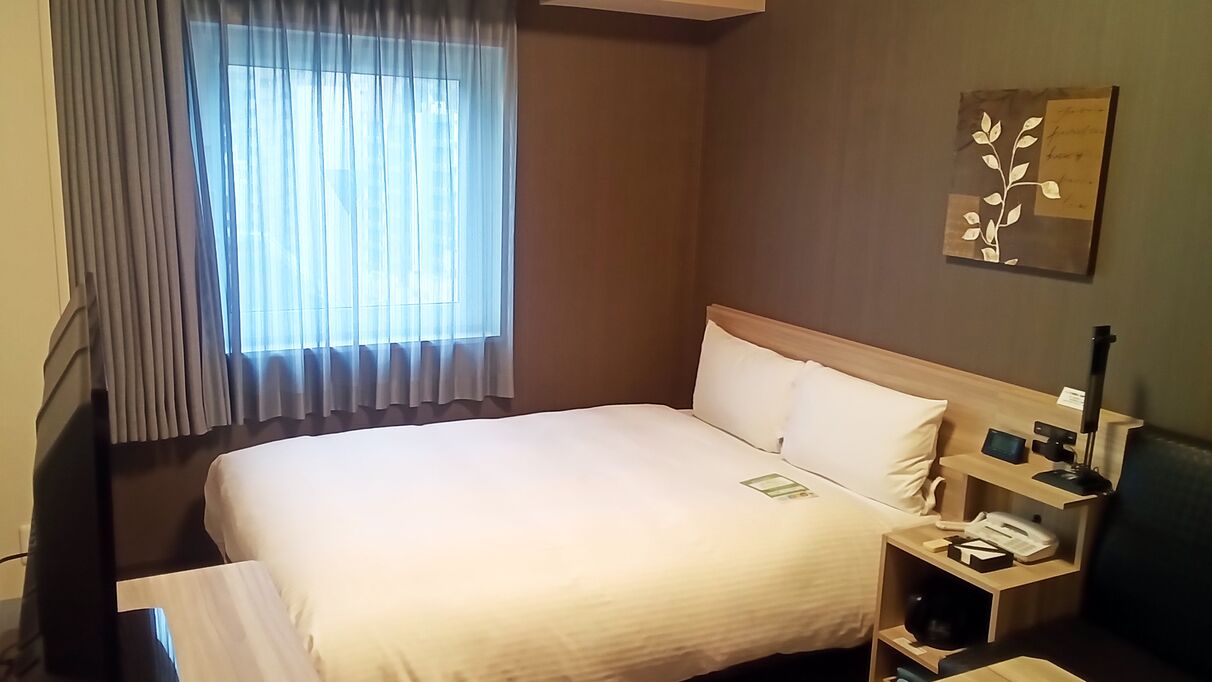 Habitació d'hotel amb un llit, una tauleta de nit amb una làmpada, un telèfon i una finestra amb cortines.